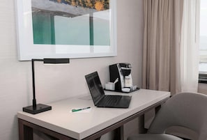 A hotel room with a desk, a laptop, a coffee maker, a framed picture, and a chair.