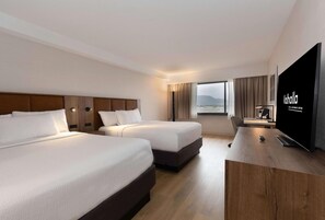 A hotel room with two beds, a TV, a desk, and a window with a view.