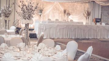 Un salón de banquetes con mesas redondas dispuestas para un evento formal, cubiertas con tela blanca e iluminadas con luces de cadena.