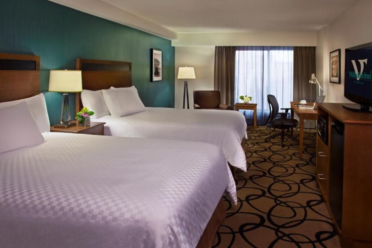 Quarto de hotel com duas camas, uma escrivaninha, uma cadeira, televisão e janela com cortinas.