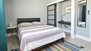Ein Schlafzimmer mit Bett, Deckenventilator, Nachttisch, kleinem Tisch mit Lampe, Spiegel und einer Schiebetür zu einem anderen Raum.