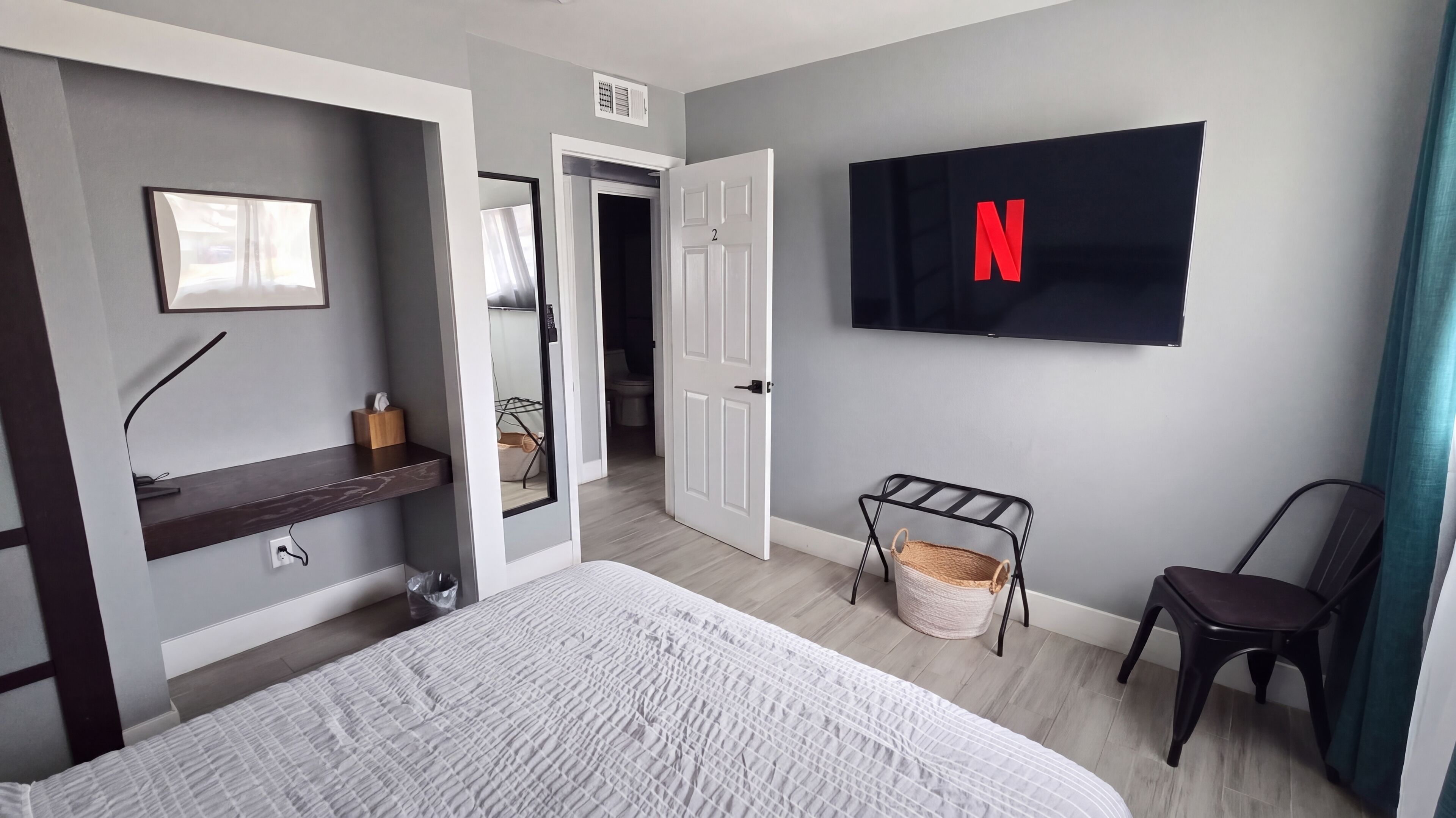 A modern bedroom with a bed, a mounted TV displaying the Netflix logo, a chair, and a small shelf with a tissue box.