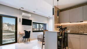 A modern kitchen with a built-in island, a flat-screen TV mounted on the wall, and a large window with a view of the city. - Royalty Suites Psyrri (Athens)