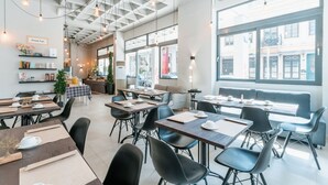 A modern dining area with tables, chairs, and a large window offering a view of the street. - Royalty Suites Psyrri (Athens)