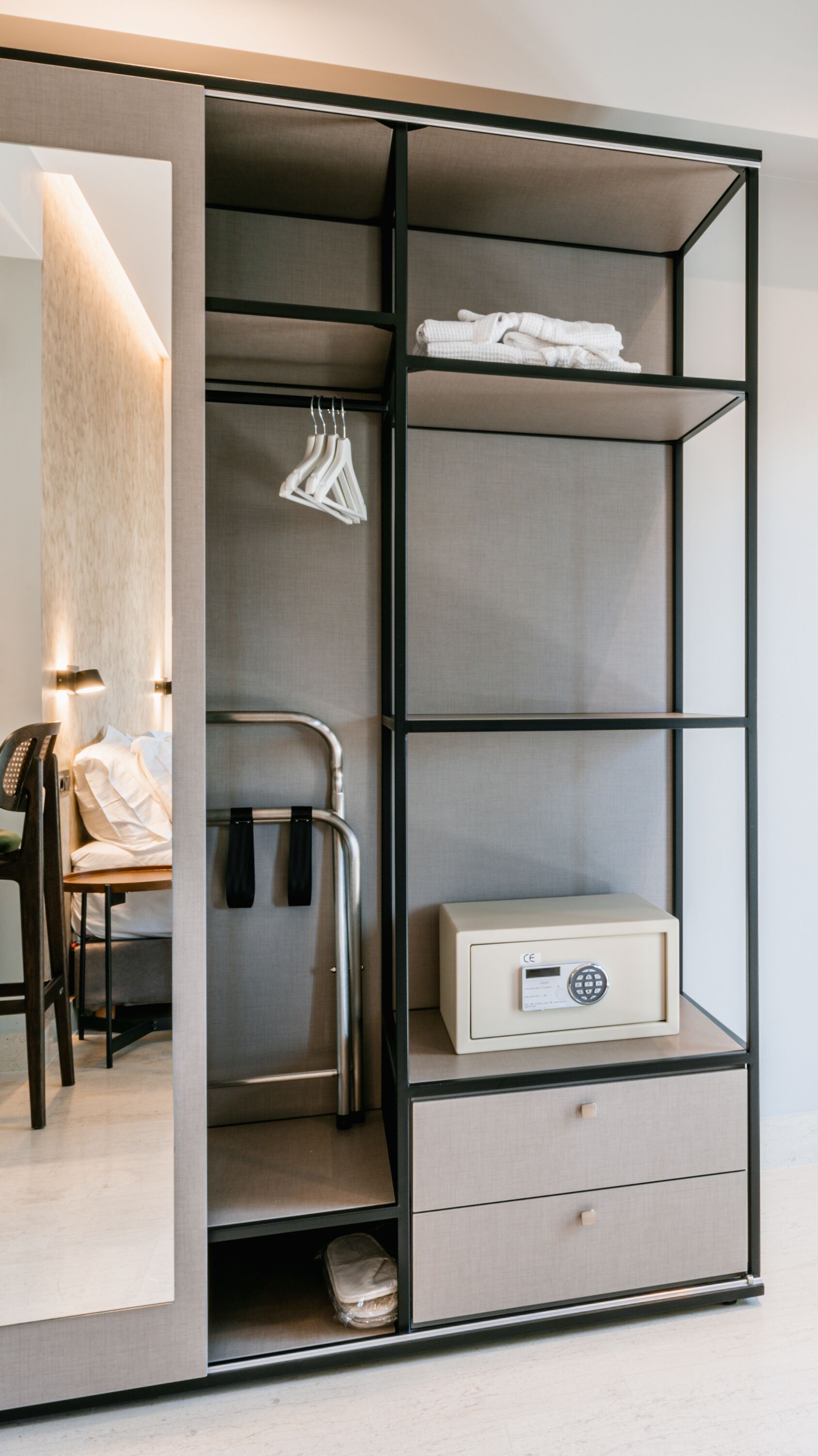 Una habitación de hotel moderna con armario empotrado, mesita de noche, una silla, un escritorio, lámpara de escritorio, teléfono, perchero, maleta y una caja fuerte.