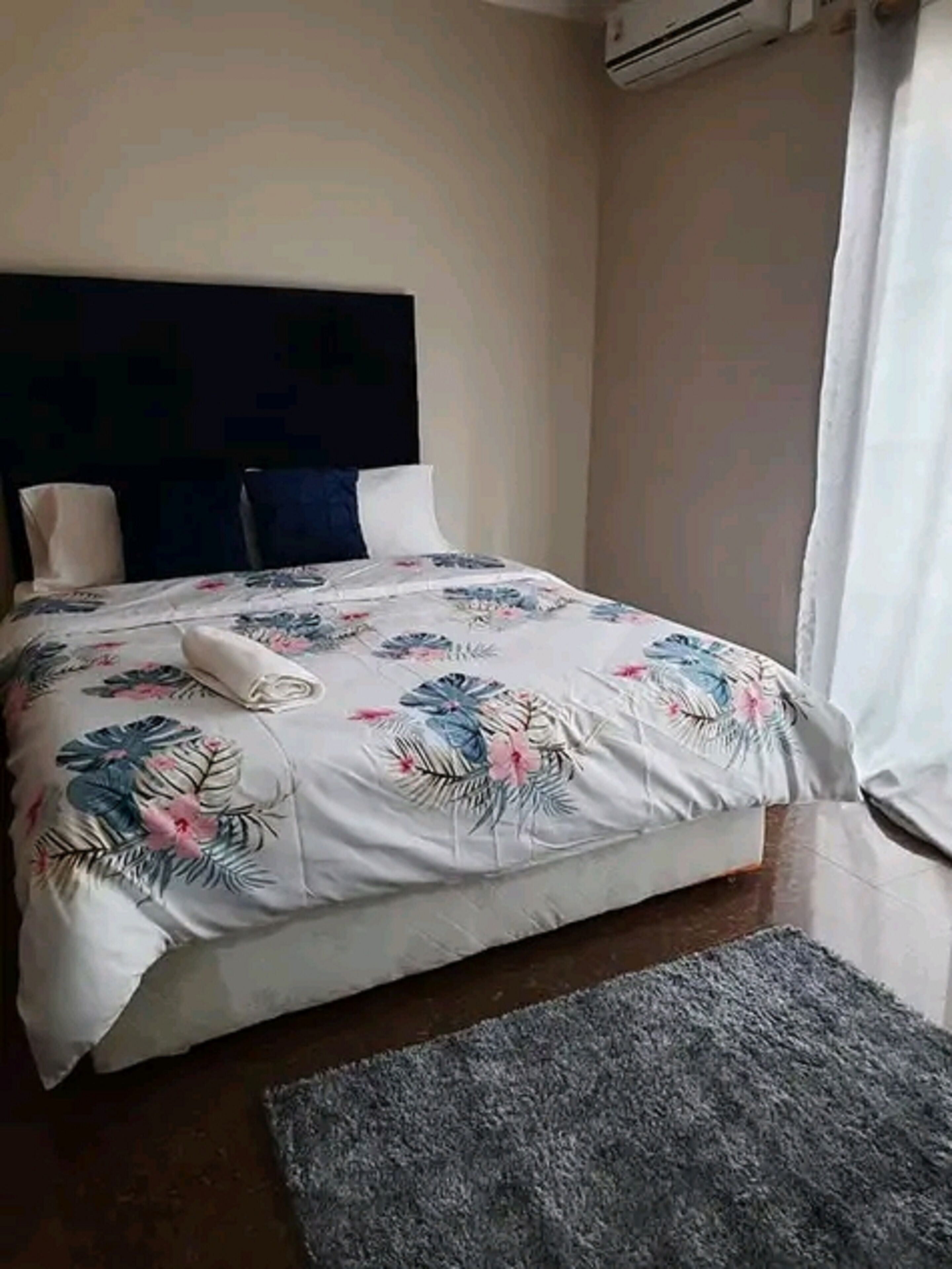 Una cama bien hecha con ropa de cama floral, cabecera oscura y alfombra gris sobre suelo de baldosas.