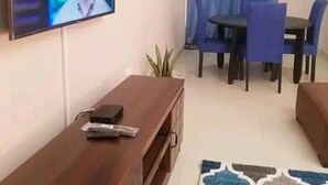 A living room with a TV mounted on the wall, a wooden entertainment unit, a dining area with blue chairs, and a patterned rug.