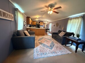 Ein Wohnzimmer mit zwei Sofas, einem Couchtisch, einem Teppich, einem Deckenventilator und einer Küche im Hintergrund.