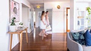 A hallway with a wooden floor, a white sofa with floral cushions, and a white wall with a framed picture. - The Otto at Moffat Beach (Moffat Beach)
