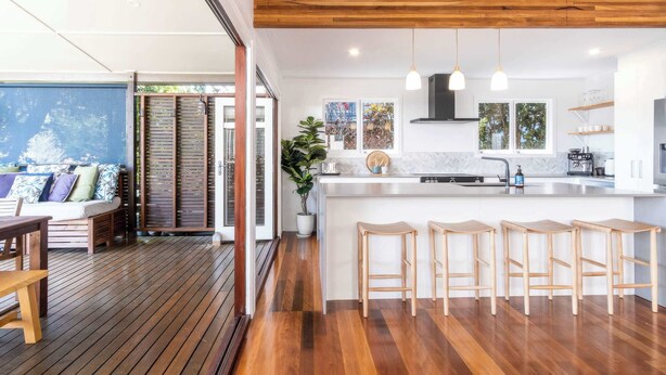 A modern kitchen with wooden flooring, a central island with stools, pendant lighting, and a view of the outdoors. - The Otto at Moffat Beach (Moffat Beach)