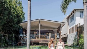 A swimming pool with two people sitting on the edge, a covered patio area, and a modern house with a staircase. - The Otto at Moffat Beach (Moffat Beach)