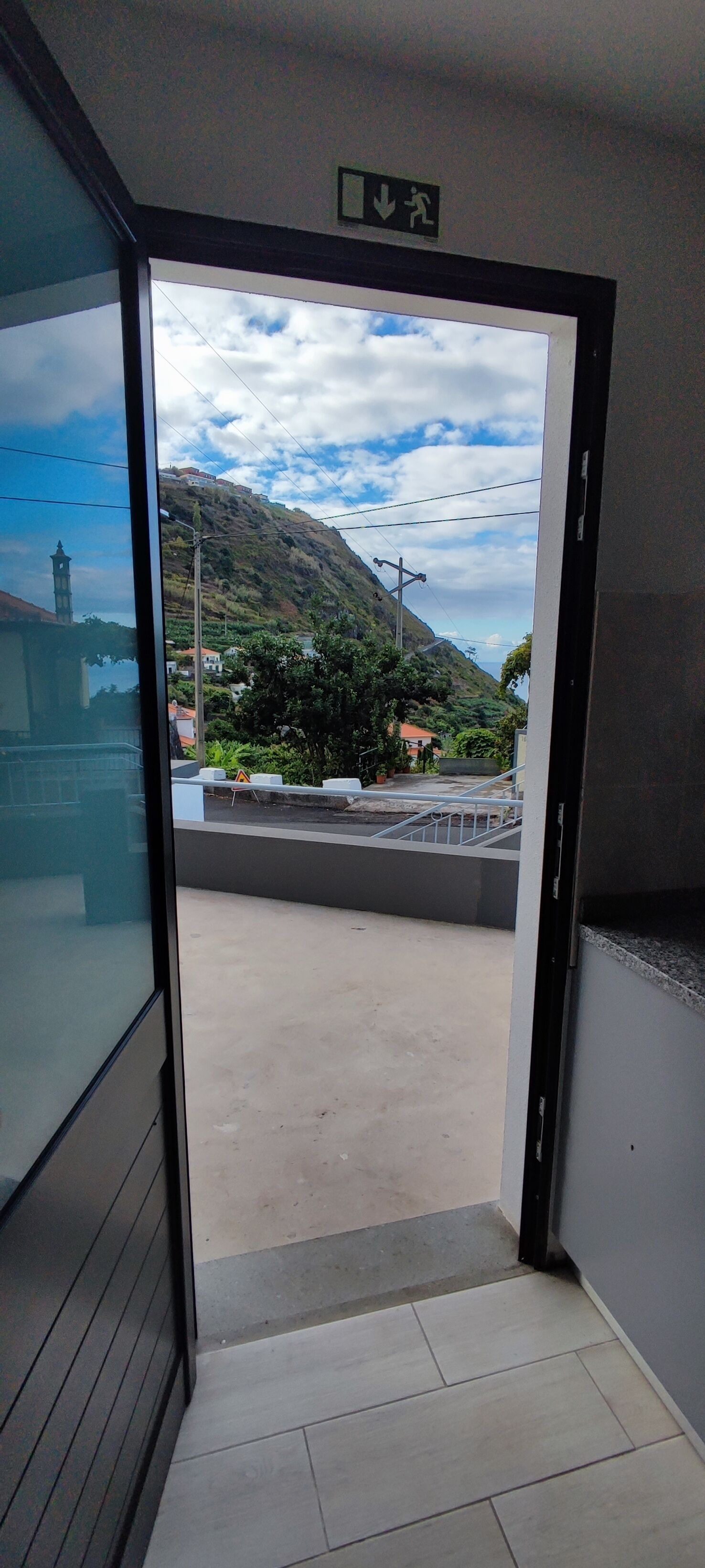An open door leading to a balcony with a view of a hillside, a staircase, and a small building in the distance.