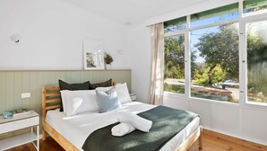 A bedroom with a bed, a bedside table, a window with a view of trees, and a wall-mounted picture. - Anglesea Beach House 2 (Anglesea)