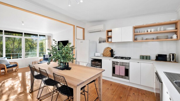 A modern kitchen with white cabinetry, a wooden dining table, and a large window overlooking greenery. - Anglesea Beach House 2 (Anglesea)