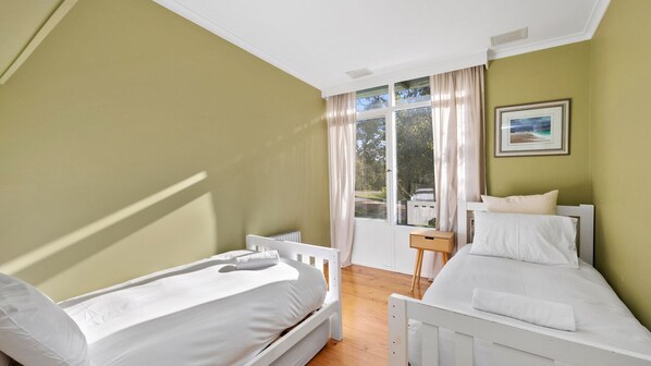 A room with two beds, a window with curtains, a small table, and a framed picture on the wall. - Anglesea Beach House 2 (Anglesea)