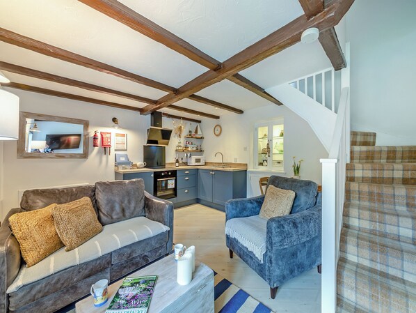 A modern living room with a sofa, armchair, and coffee table, a kitchen with a built-in oven and microwave, and a staircase with a checkered carpet.