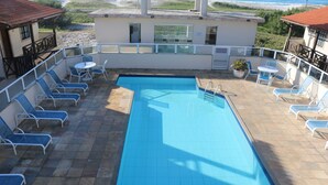 A rectangular pool with a tiled deck, surrounded by white lounge chairs and a small table. - CASA GORETE PRAIA DAS DUNAS RESIDENCE CLUB (Cabo Frio)