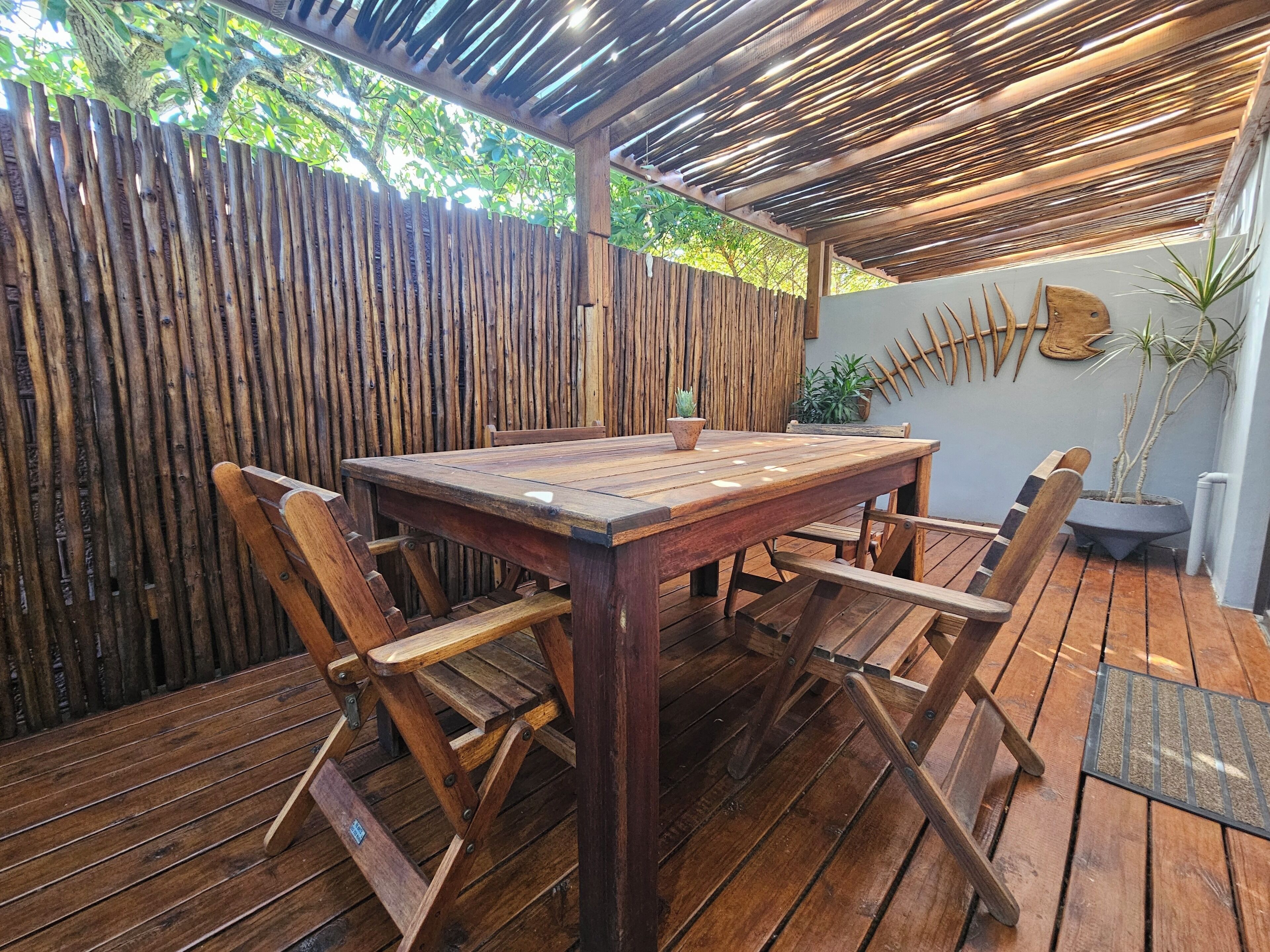 Une terrasse extérieure en bois, avec une table et des chaises, entourée d’une clôture en bambou et agrémentée de plantes en pot.
