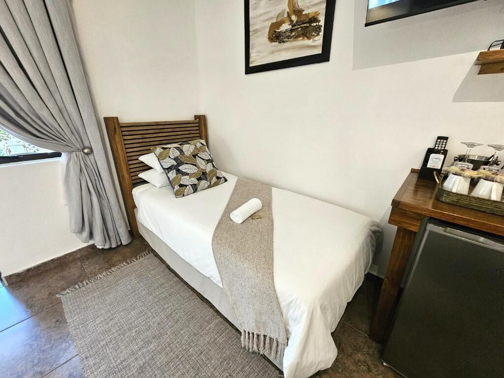 Una cama individual con cabecera de madera, una manta gris y un cojín con estampado de hojas. Una mesita de madera con tetera y tazas está junto a la cama.