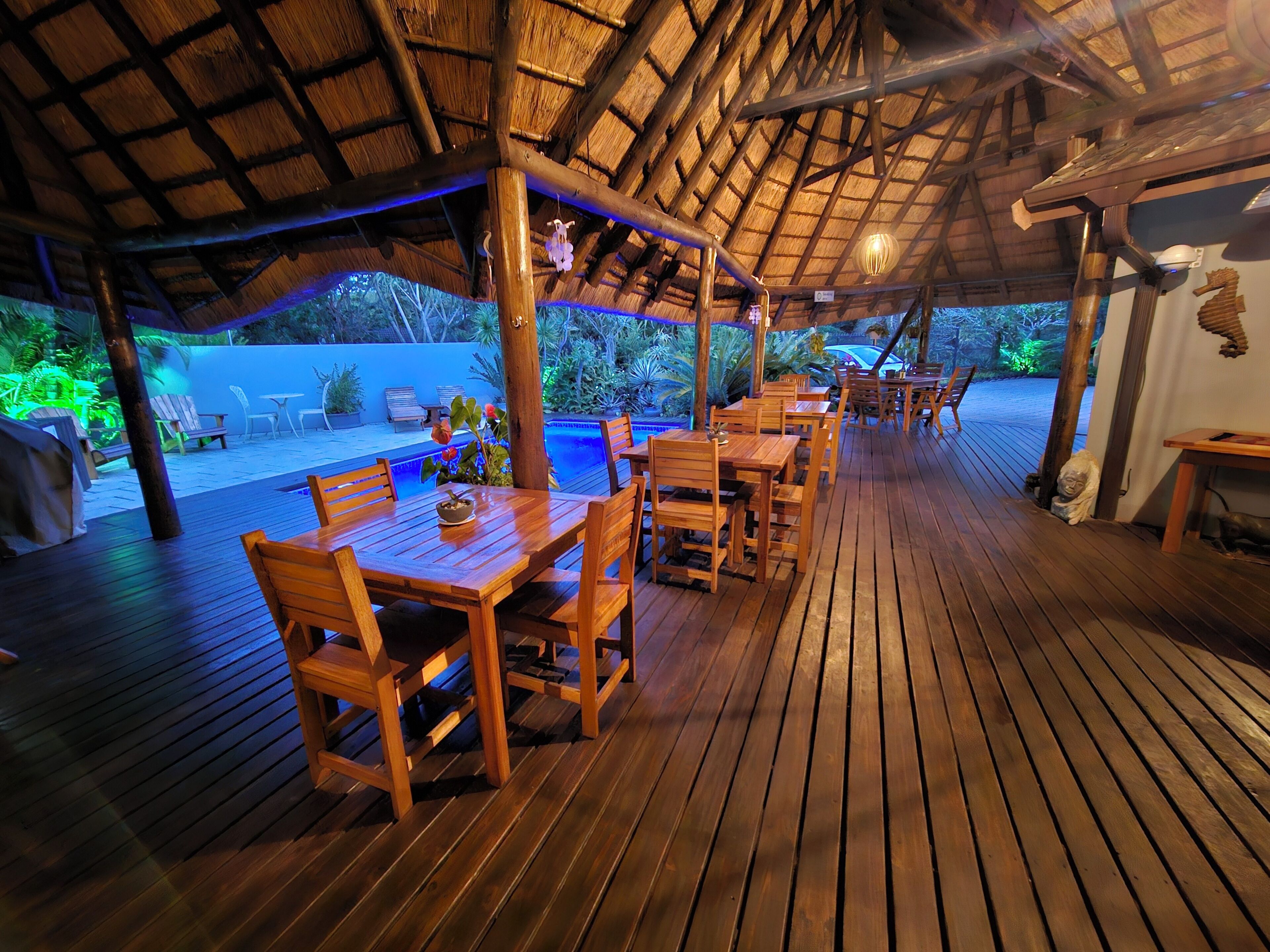 An outdoor wooden deck with tables and chairs, a thatched roof, and a pool area.