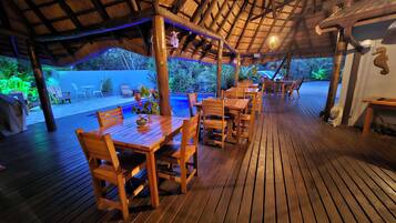 An outdoor wooden deck with tables and chairs, a thatched roof, and a pool area.