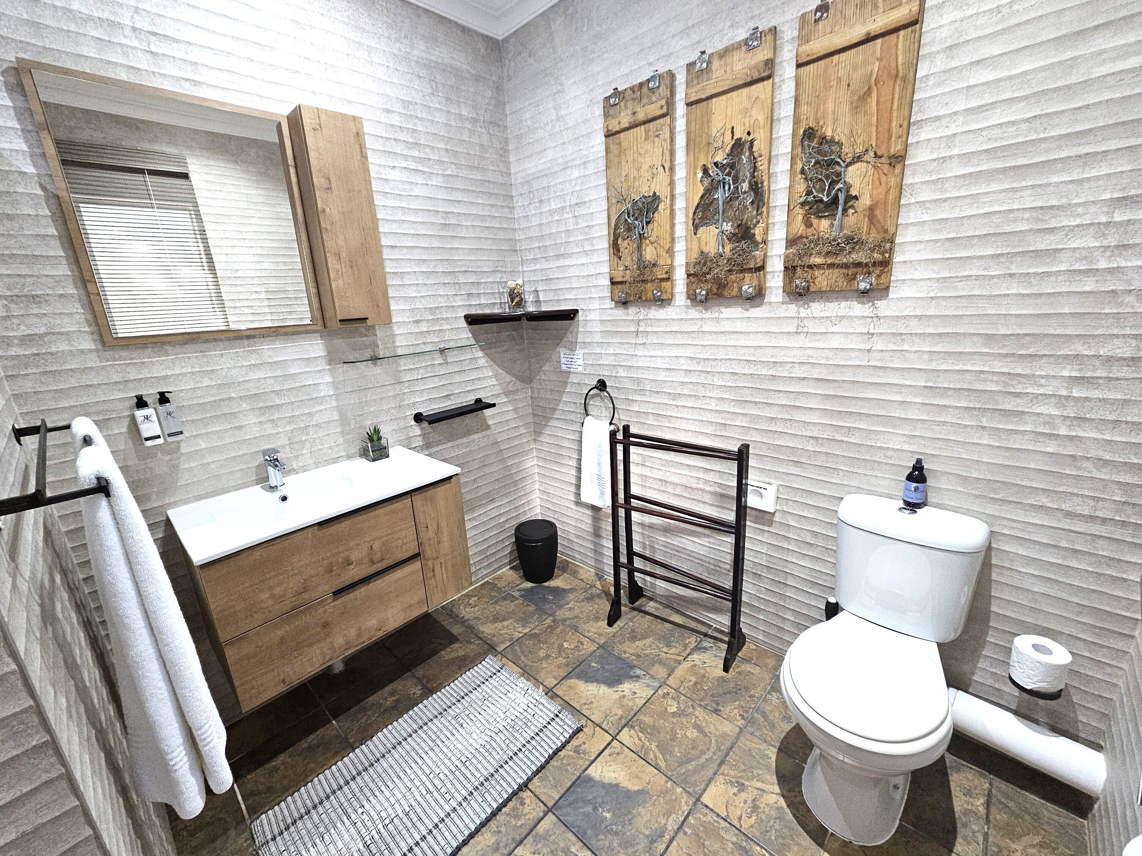 Une salle de bain avec un meuble-lavabo en bois, un lavabo blanc et des toilettes. On y trouve un porte-serviettes, une petite étagère et un élément décoratif mural au-dessus du lavabo.