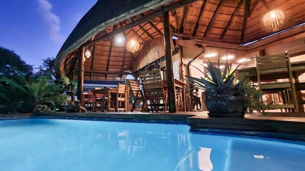 Un área junto a la piscina con una estructura de techo de paja, muebles de madera y una planta grande en maceta.