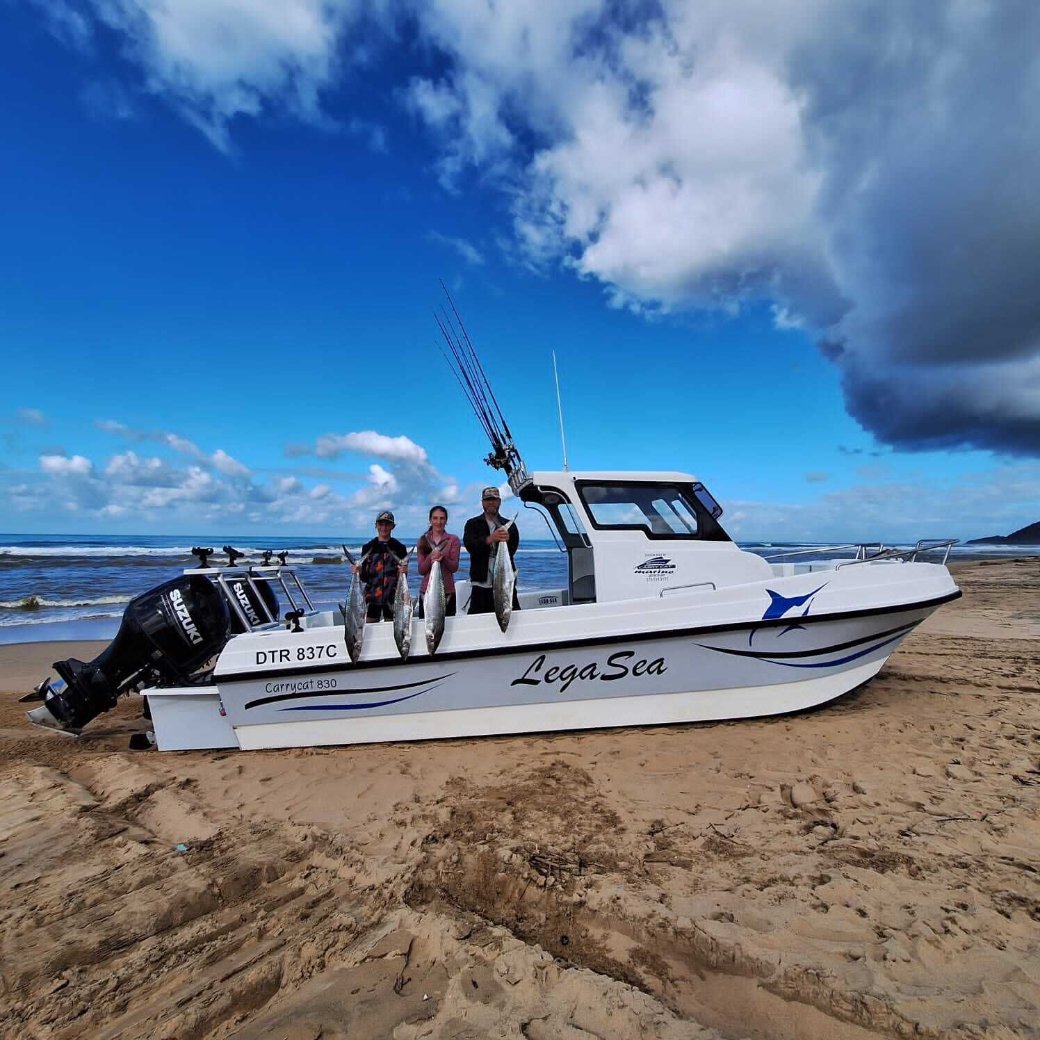 A boat named 'LegaSea' with a Suzuki outboard motor, docked on a sandy beach with the ocean and a partly cloudy sky in the background.