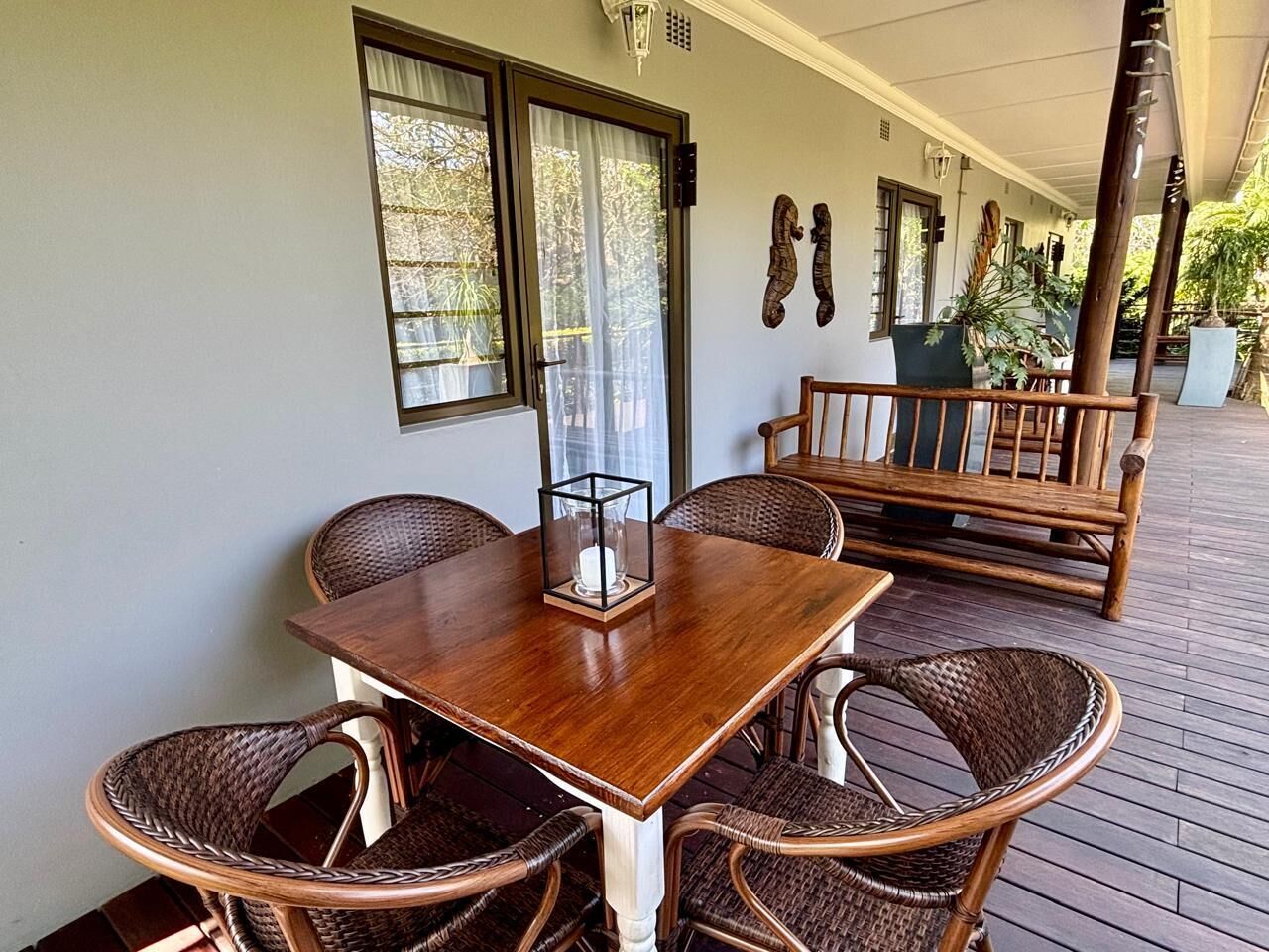 A covered outdoor seating area with a wooden table and wicker chairs, a bench, and potted plants.