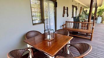 A covered outdoor seating area with a wooden table and wicker chairs, a bench, and potted plants.