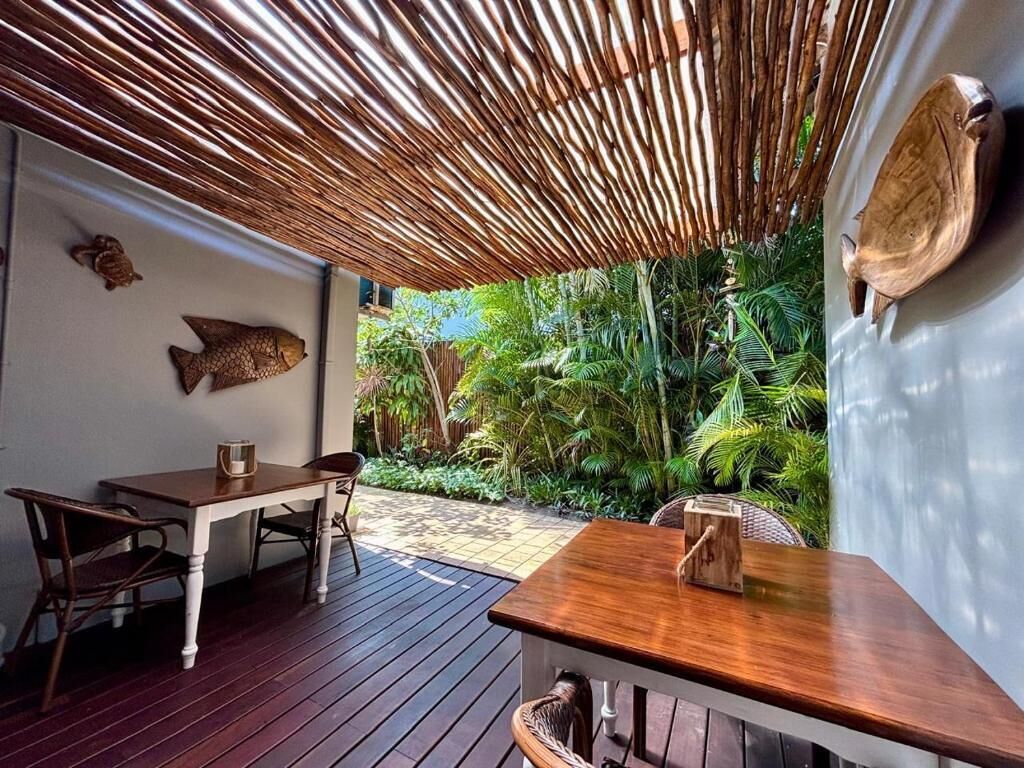 Um pátio externo com mesa e cadeiras de madeira, teto de palha e plantas tropicais.