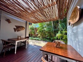 Un patio al aire libre con mesa y sillas de madera, techo tejido y plantas tropicales.