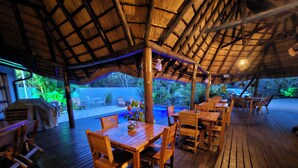 Una terraza exterior de madera con mesas y sillas, un techo de paja y un área de piscina.
