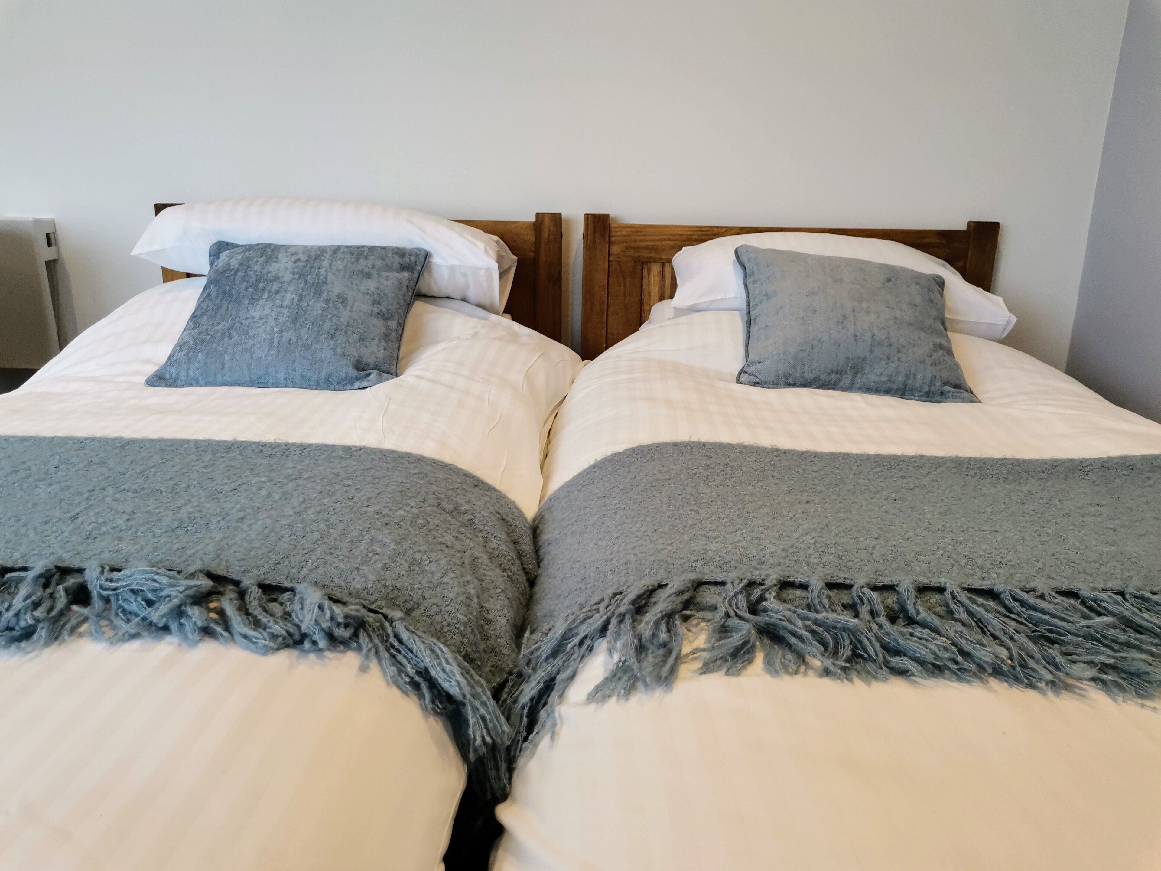 Twee eenpersoonsbedden met houten hoofdeinden, beddengoed in wit en grijs, en grijze plaids.