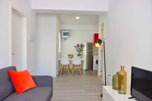 Uma sala de estar com um sofá cinza, uma almofada vermelha, uma televisão e duas garrafas de vidro sobre uma mesa branca.