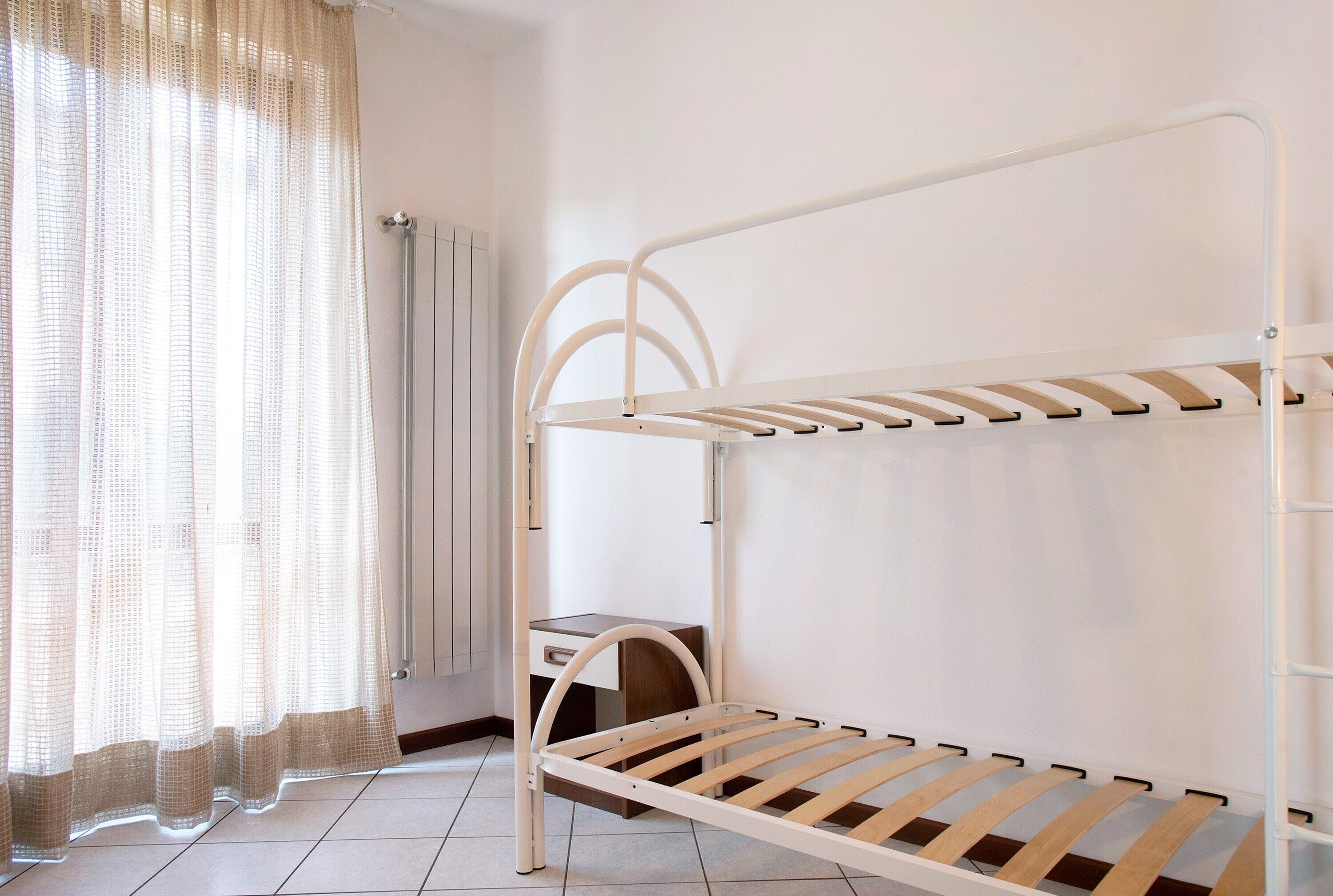 Uma cama beliche com escrivaninha embaixo, uma janela com cortinas translúcidas e um radiador.
