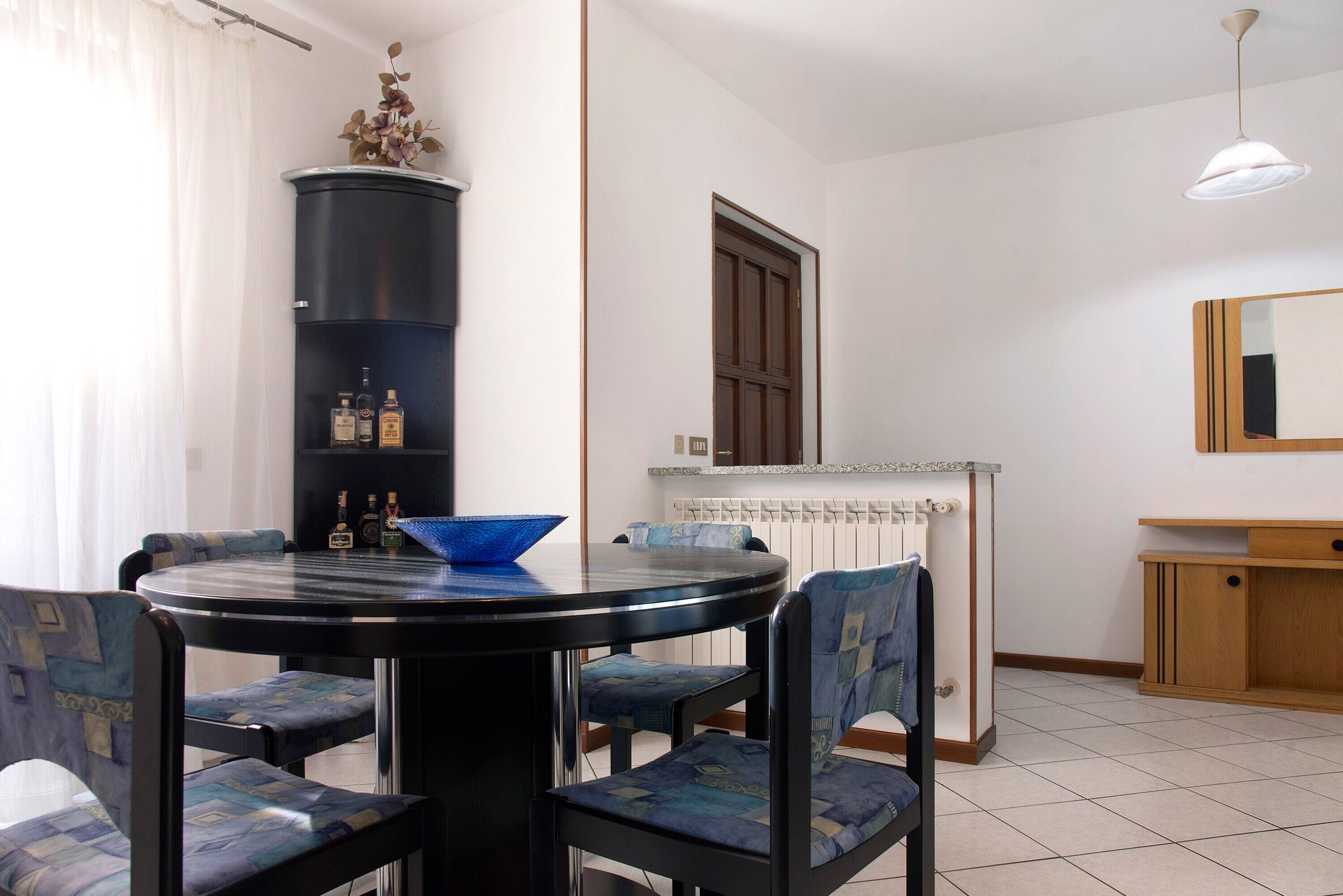 A dining area with a round table, chairs, a blue bowl, a cabinet with bottles, a radiator, and a mirror.
