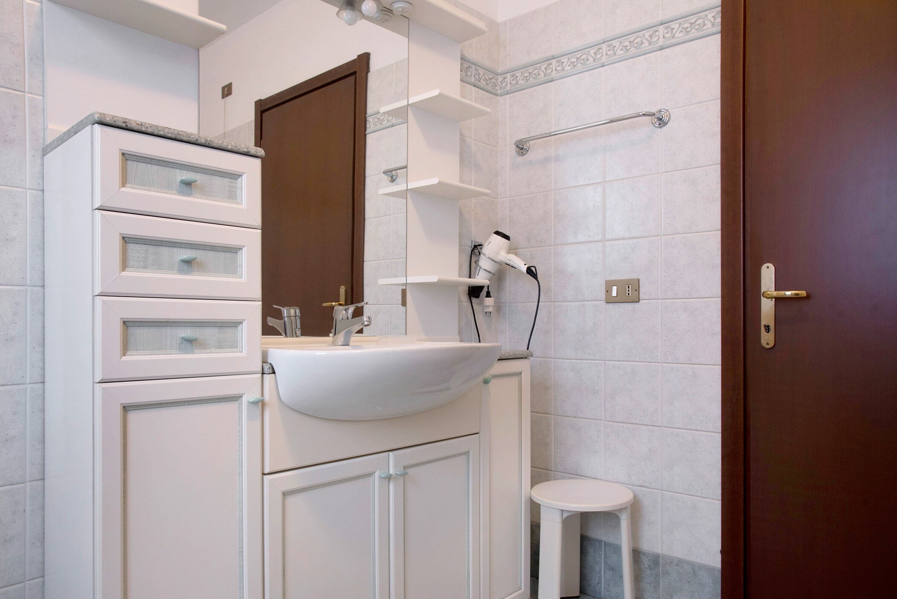 Ein Badezimmer mit Waschbecken, Spiegel und Schränken, einem Föhn und einem kleinen Hocker.