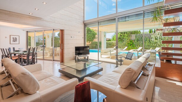 A modern living room with a glass coffee table, leather sofas, a flat-screen TV, and a view of a pool and greenery. - Villa Agusmar with Sea View, Pool & Wi-Fi (llucmajor)