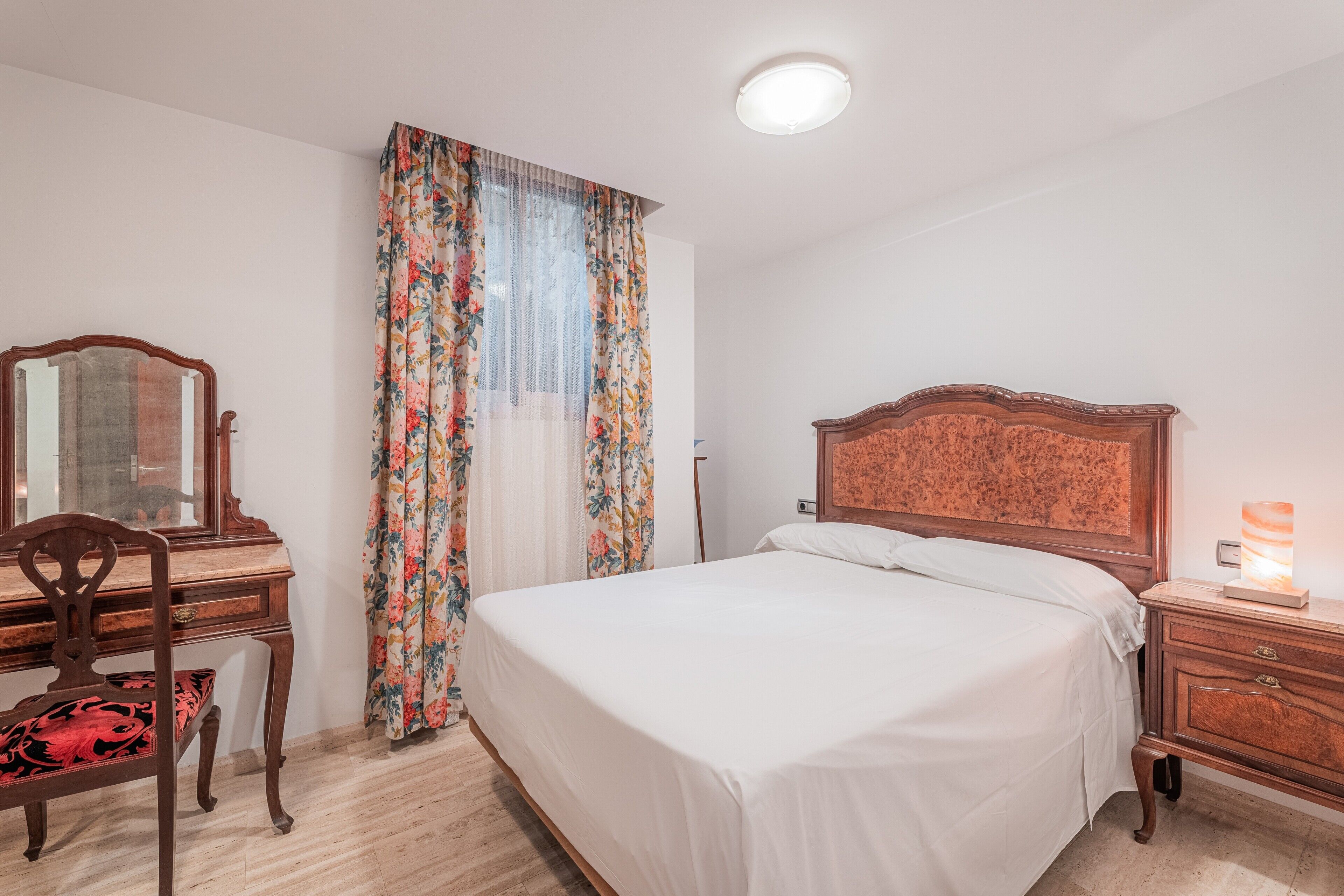 Un dormitorio con una cama de madera, una mesita de noche, un escritorio con silla y una ventana con cortinas florales.