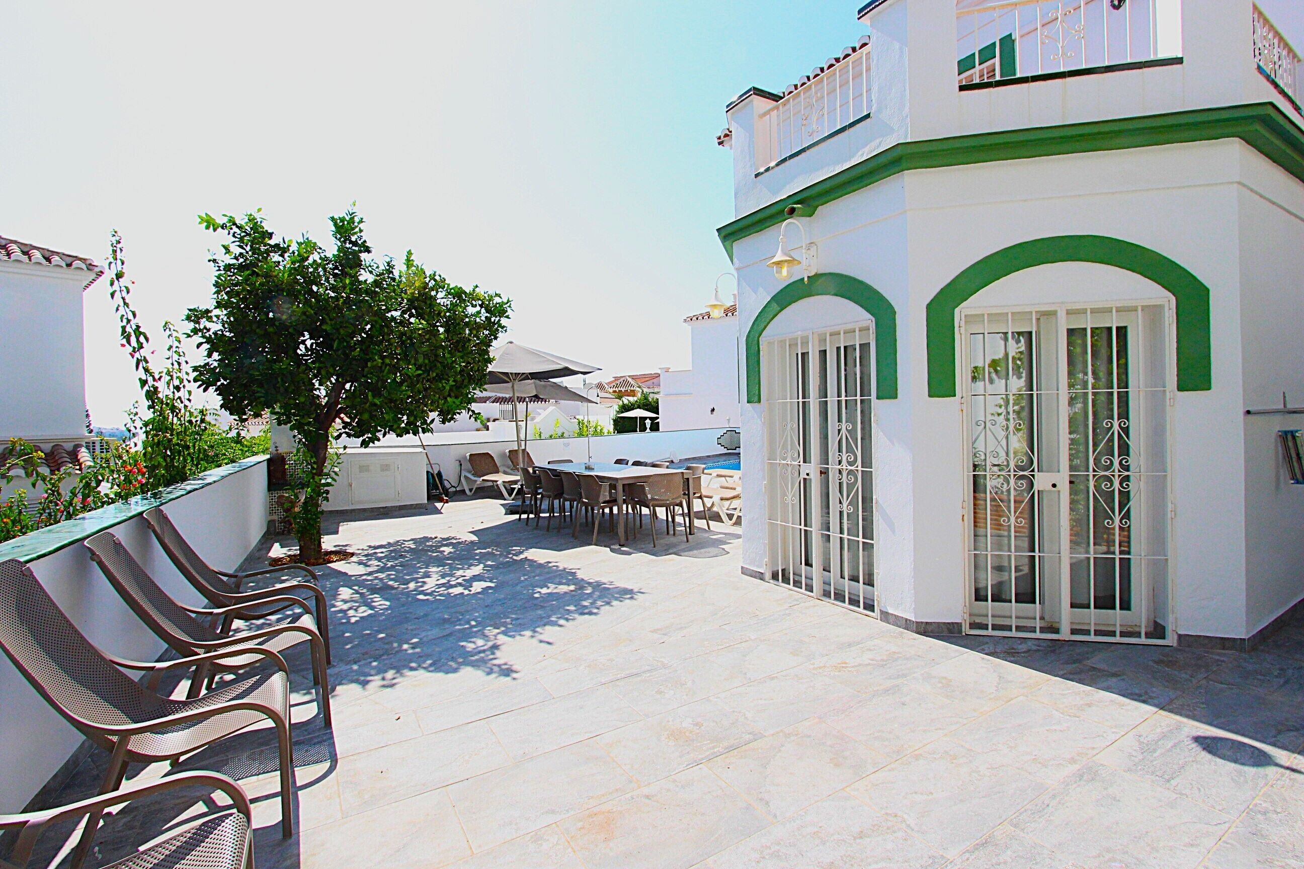 Terraza en la azotea con mobiliario exterior, piscina y un edificio con arcos verdes y paredes blancas.