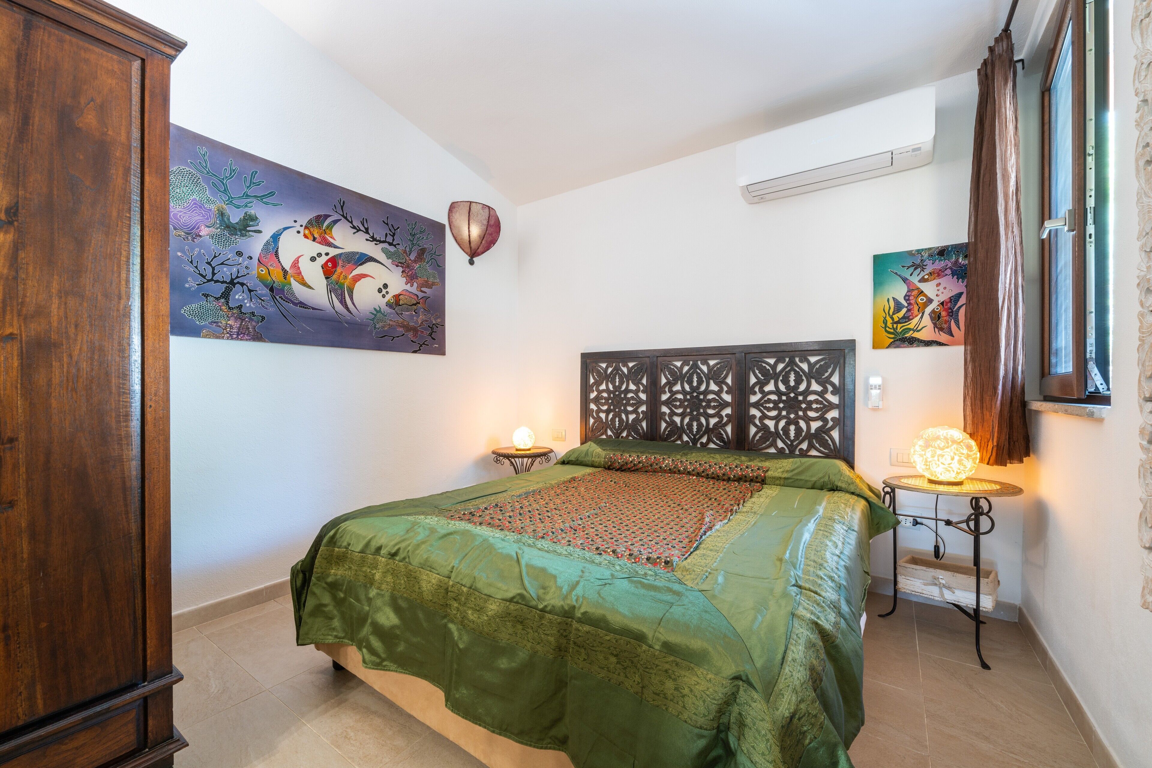 Une chambre à coucher comprenant un lit, une armoire en bois, une tête de lit décorative, une œuvre d’art accrochée au mur, une table de chevet avec une lampe et une fenêtre avec des rideaux.