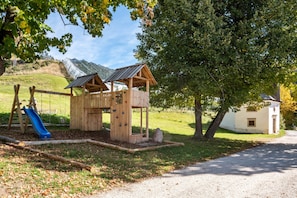 Une structure de jeux en bois, dotée d’un toboggan et d’un mur d’escalade, entourée d’arbres et d’une colline.