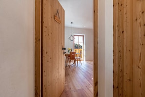 A wooden interior hallway leading to a dining area with a table and chairs, a hanging light fixture, and a window with a view.
