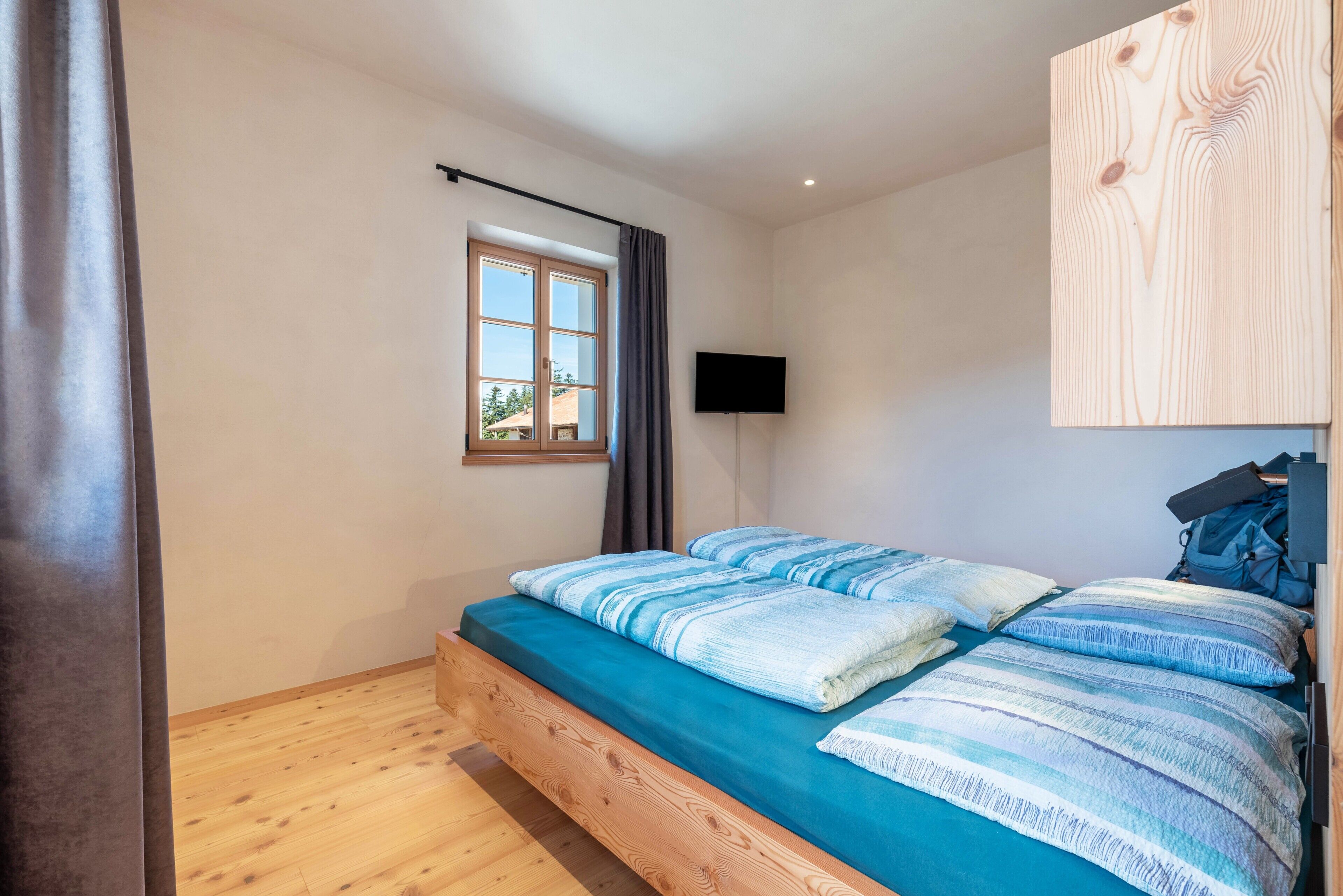 A bedroom with a wooden bed, a window with curtains, a mounted TV, and a wooden wall panel.