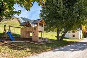A wooden play structure with a slide and climbing wall, surrounded by trees and a hill.