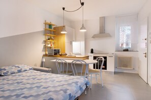 Une pièce de taille modeste comprenant un lit, une petite table à manger, des chaises, un coin cuisine et une fenêtre avec des stores.