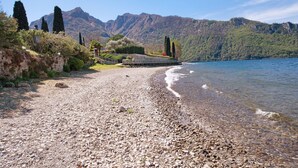 A pebbled beach with a stone wall and a small structure, overlooking a lake and mountains.