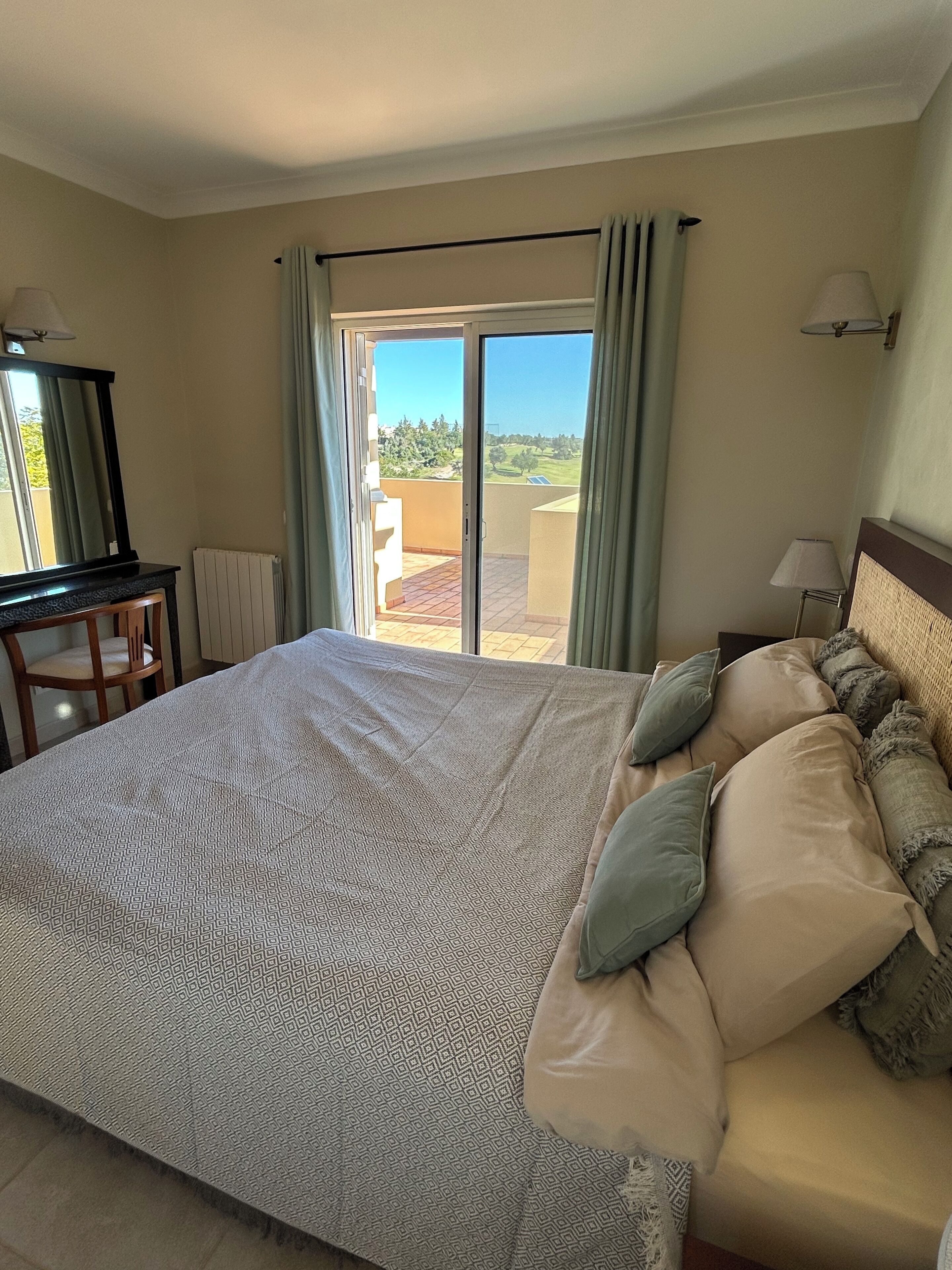 A hotel room with a large bed, a desk, a chair, and a view of the outdoors through a sliding glass door.