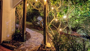 A stone pathway leading to a wooden pergola with string lights, surrounded by lush greenery and a stone wall. - Green Nest at the Hills (Guaynabo)